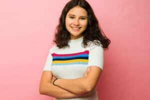 Smiling teenage girl with crossed arms on a pink background, reflecting confidence after rhinoplasty